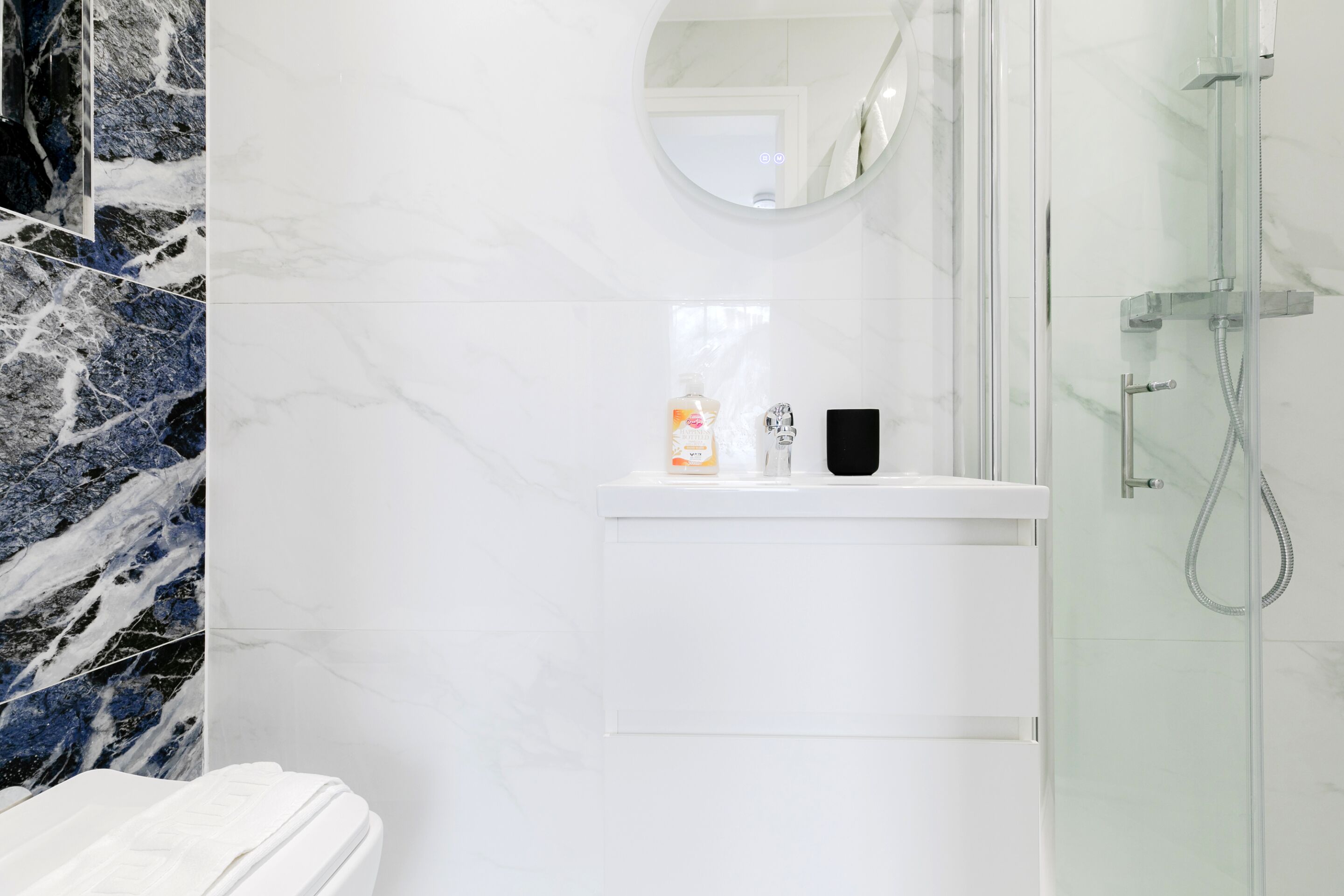 A bathroom with a marble wall, a glass shower enclosure, and a white sink with toiletries.