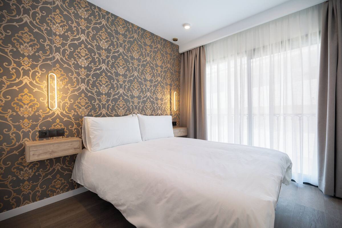 Un lit bien fait, avec du linge de lit blanc, une table de chevet en bois et un mur recouvert d’un papier peint à motifs.