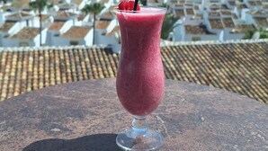 Un batido rosa en un vaso con pajita negra, sobre una superficie de piedra con vista a un pueblo costero al fondo.