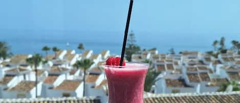 A pink smoothie in a glass with a black straw, placed on a stone surface with a view of a coastal town in the background.