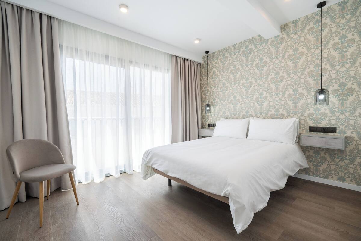 Une chambre à coucher avec un lit, une chaise, une fenêtre avec des rideaux et un mur recouvert d’un papier peint à motifs.