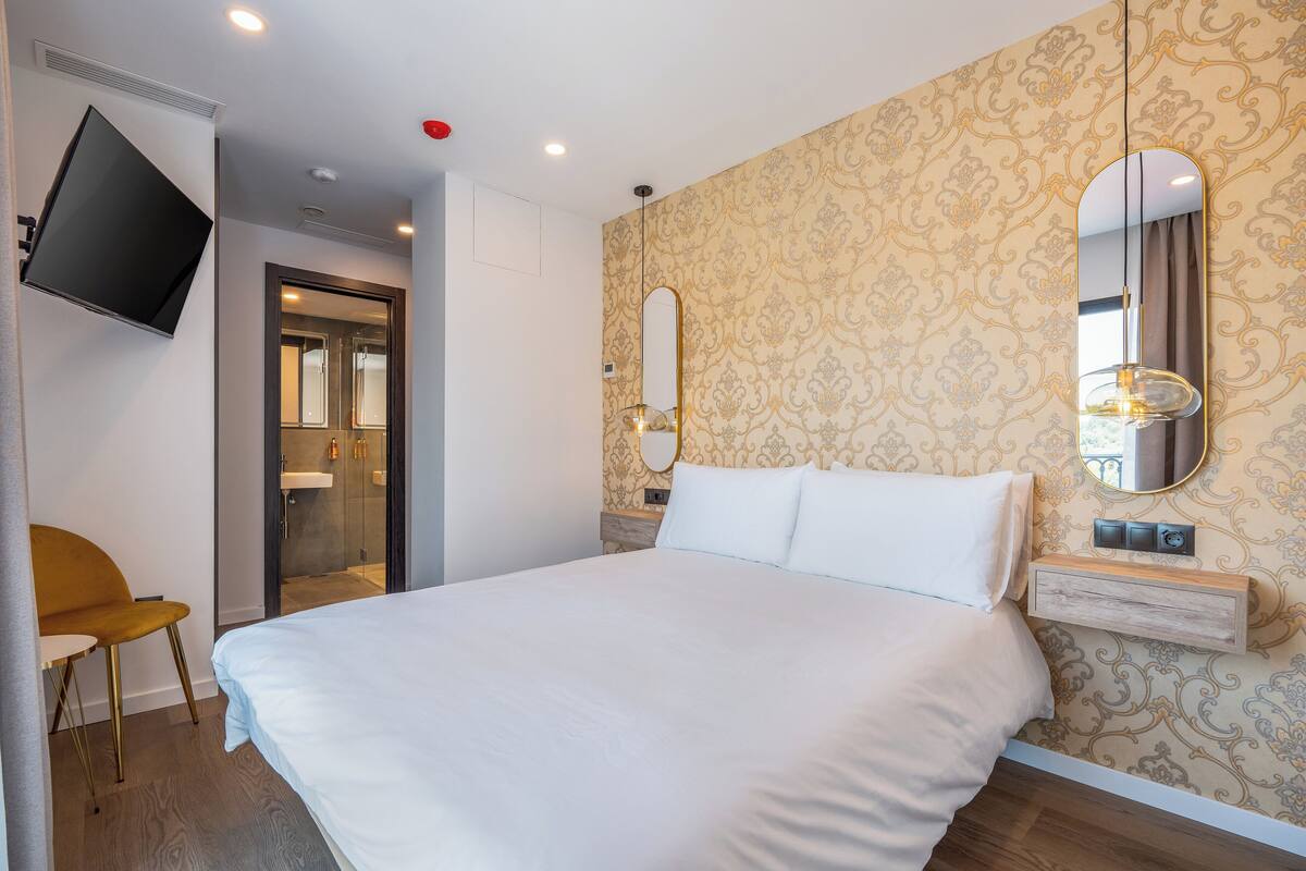 Une chambre d’hôtel moderne avec un grand lit, un téléviseur fixé au mur, un miroir et une petite table de chevet.