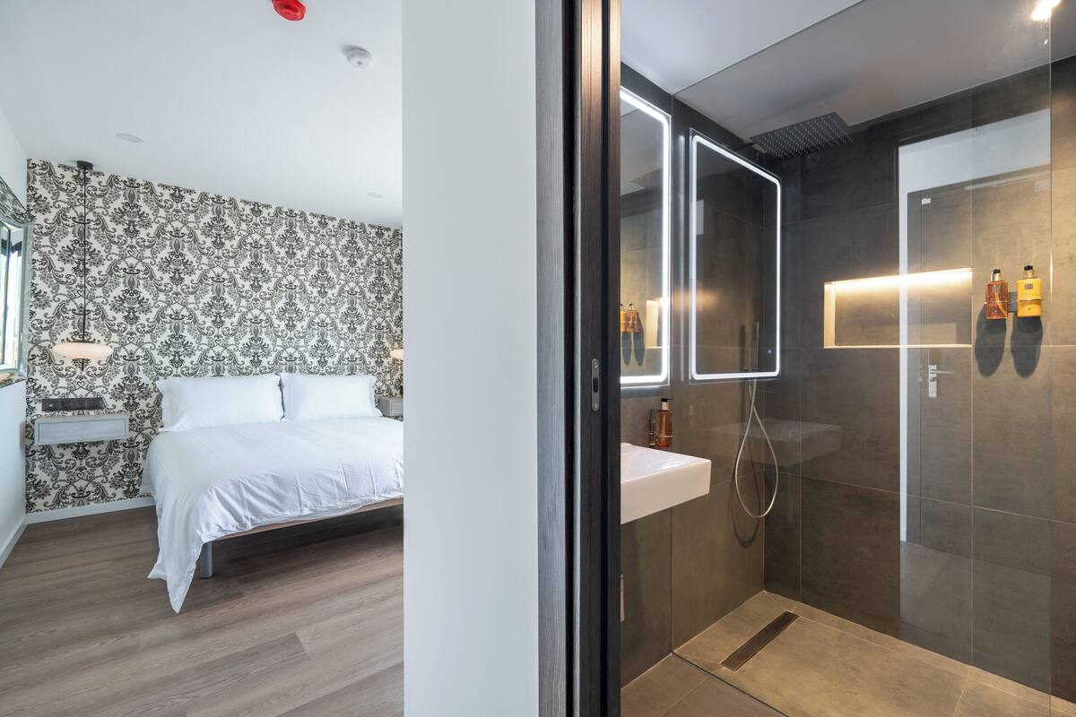 Une chambre à coucher moderne, dotée d’un grand lit, d’une table de chevet et d’un papier peint à motifs. À côté se trouve une salle de bain avec une douche en verre, un lavabo et un miroir.