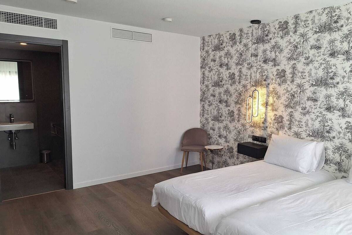 Une chambre à coucher comprenant un lit, une table de chevet, une chaise, une petite table et une salle de bain avec un lavabo et un miroir.