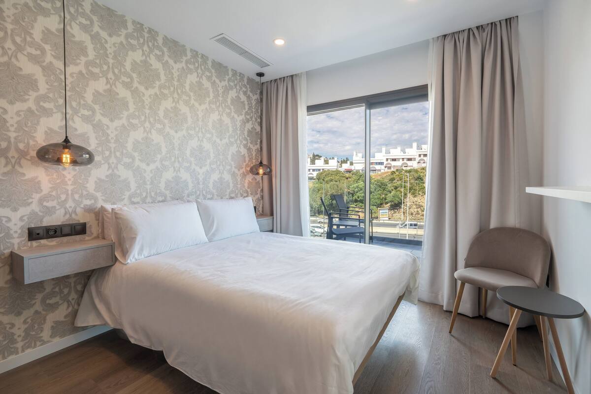 Une chambre d’hôtel moderne, dotée d’un grand lit, d’une chaise, d’une petite table et offrant une vue sur la ville par la fenêtre.