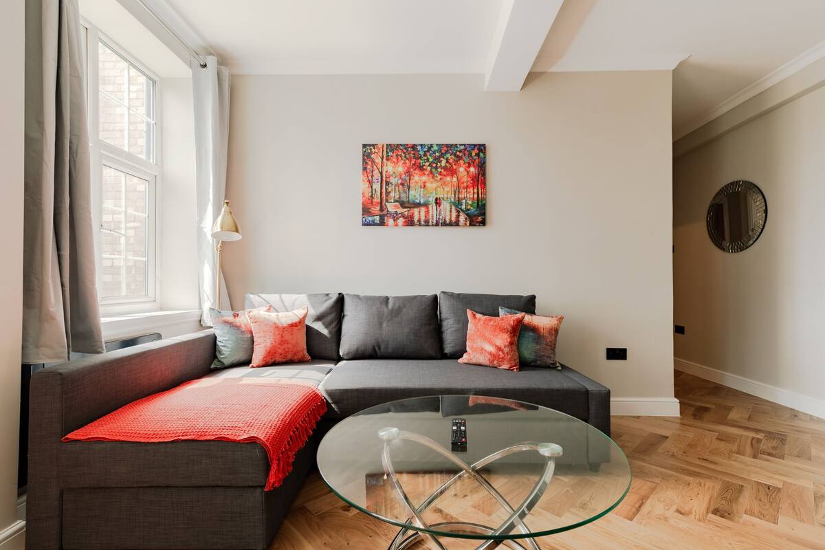 A modern living room with a grey sofa, red throw pillows, a glass coffee table, and a painting of a forest scene.