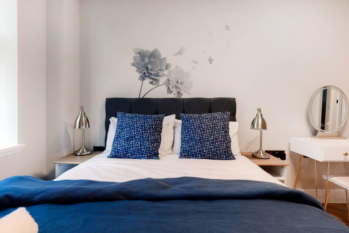 A neatly made bed with a dark blue comforter and two patterned pillows, a bedside table with a lamp, and a dresser with a mirror.