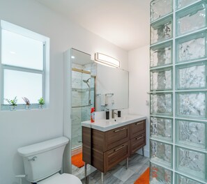 Un baño moderno con cabina de ducha de vidrio, inodoro blanco, tocador de madera con espejo y ventana sobre el lavabo.
