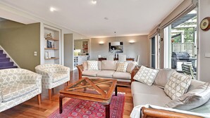 A modern living room with a sofa, armchairs, and a coffee table. There is a kitchen area in the background with a dining table and chairs. - Villa 3br Gruner Veltliner located within Cypress Lakes Resort (Pokolbin)
