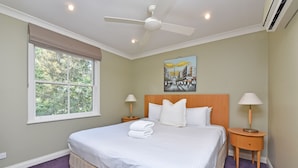 A hotel room with a bed, two bedside tables with lamps, a ceiling fan, a window with a view of trees, and a painting on the wall. - Villa 3br Gruner Veltliner located within Cypress Lakes Resort (Pokolbin)