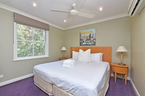 A hotel room with a bed, two bedside tables with lamps, a ceiling fan, a window with a view of trees, and a painting on the wall.