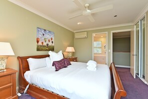 A hotel room with a bed, a ceiling fan, a painting, an air conditioning unit, a nightstand, a lamp, a bathroom door, and a towel stack.