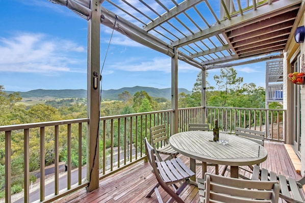 A wooden deck with a round table and chairs, overlooking a scenic landscape with hills and trees.