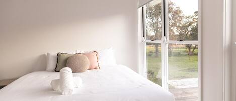 Un dormitorio con un ventanal grande, una cama con ropa de cama blanca y un perro blanco pequeño.