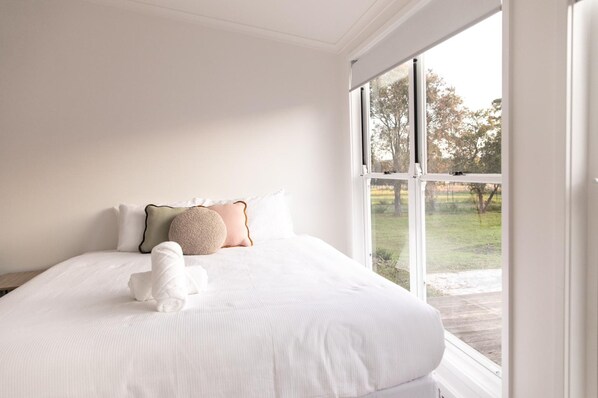 A bedroom with a large window, a bed with white bedding, and a small white dog.
