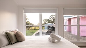 A bedroom with a large bed, a view of a garden, and a pink building outside. - Heart of Lovedale (Keinbah)