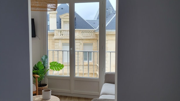 Un balcón con vistas a edificios, una lámpara colgante de mimbre, una mesa redonda con una planta y una cama.