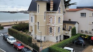 Un edificio de varios pisos con una torre destacada, un estacionamiento con varios autos y vista al mar de fondo.