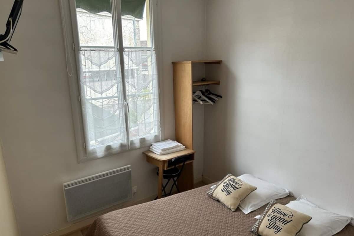 Een slaapkamer met een bed, een bureau, een stoel, een raam met jaloezieën en een plank met hangende spullen.