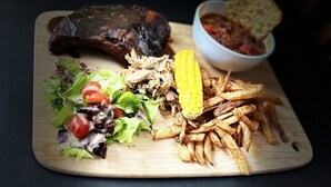 Un repas composé d’un steak, d’une salade, d’épis de maïs, de frites et d’un bol de chili, le tout servi sur une planche en bois.