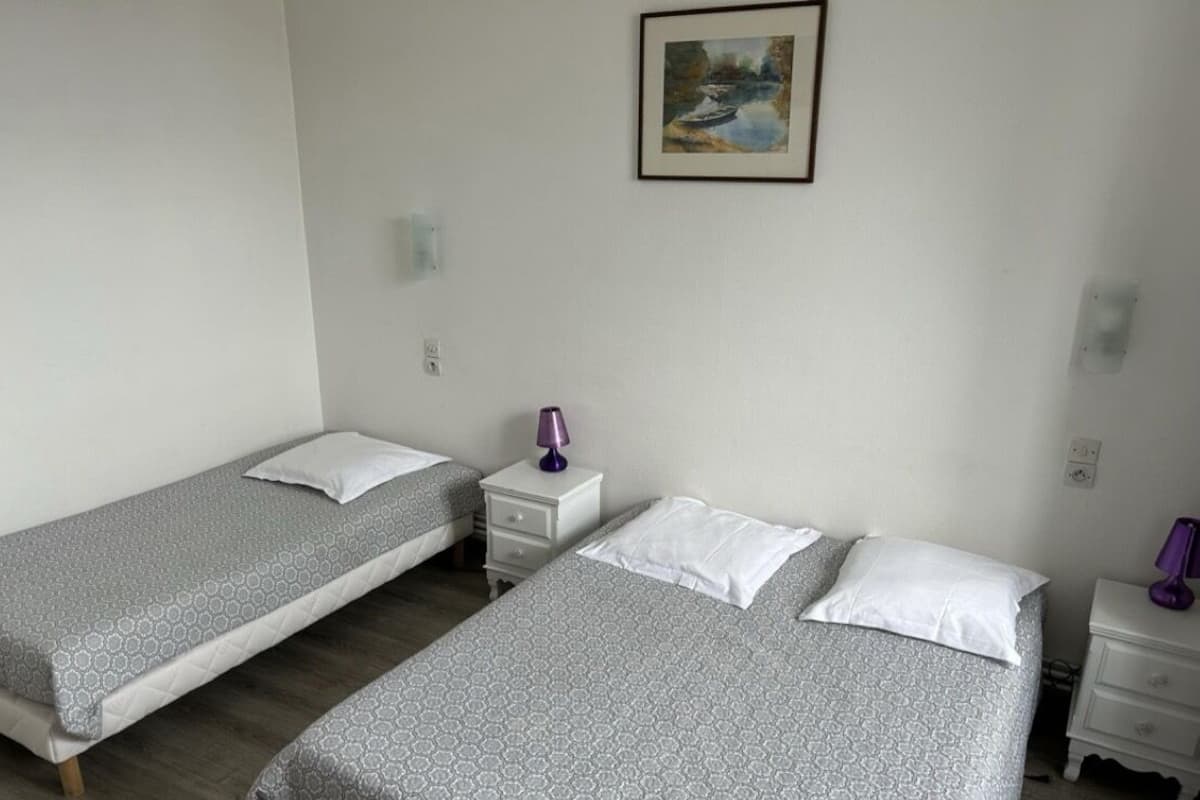 Een kamer met twee eenpersoonsbedden, elk voorzien van een wit kussen en een bed met een patroon. Tussen de bedden staat een klein wit nachtkastje met een paarse lamp. Daarboven hangt een schilderij in een lijst aan de muur.