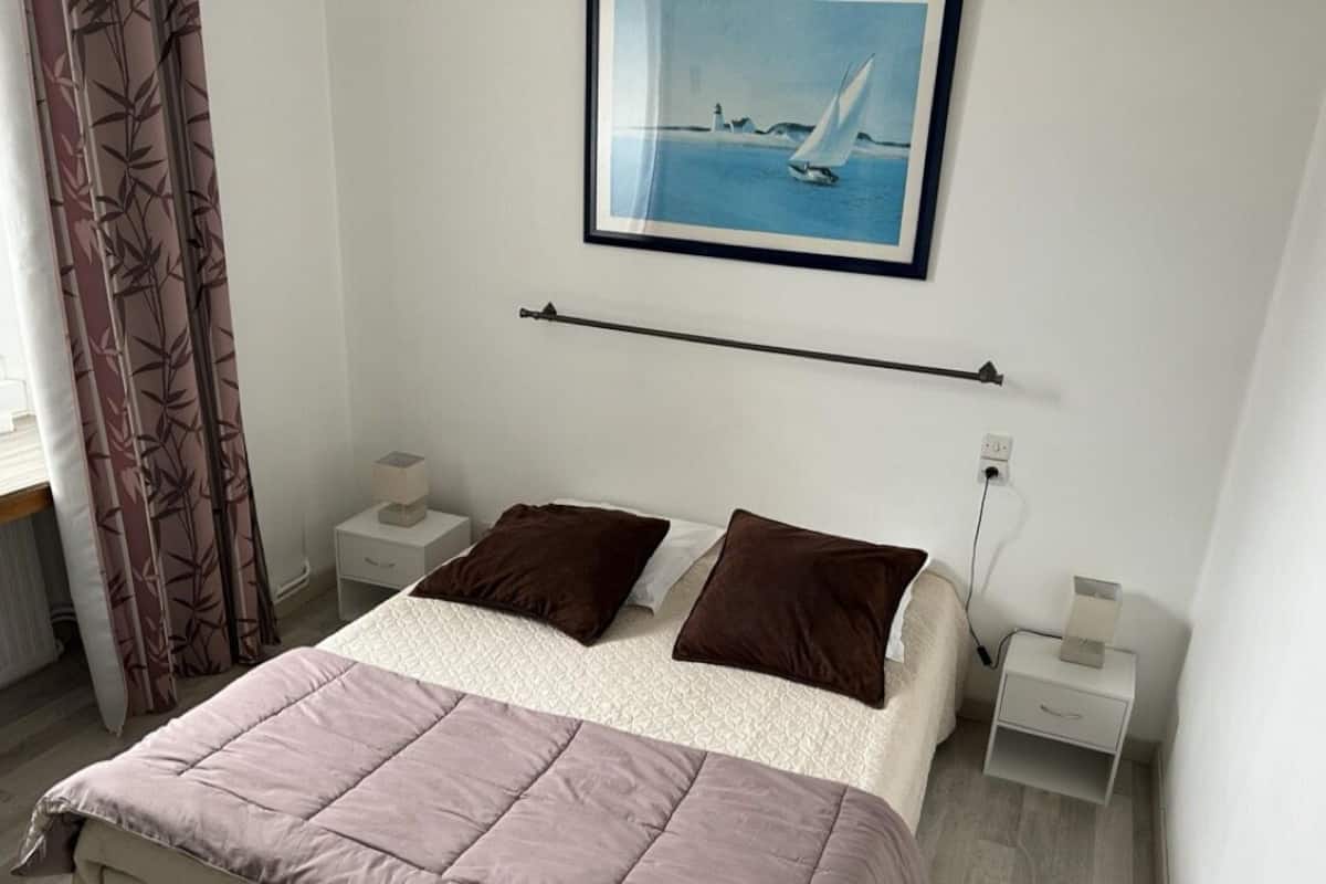 Een slaapkamer met een bed, twee kussens, een nachtkastje, een lamp, een foto aan de muur en een gordijn.