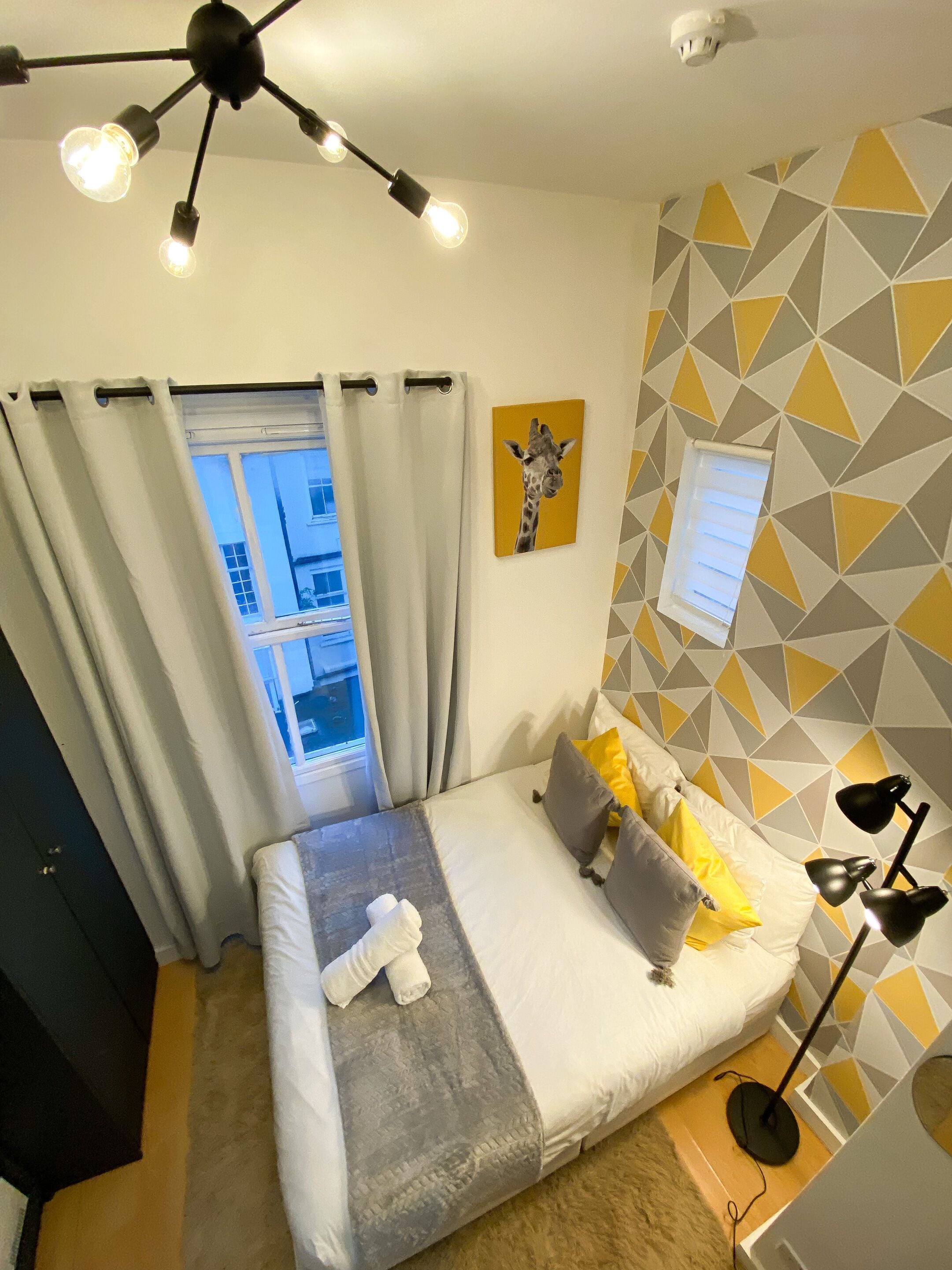 A hotel room with a bed, a window with curtains, a wall with geometric patterns, a painting of a giraffe, and a ceiling light fixture.