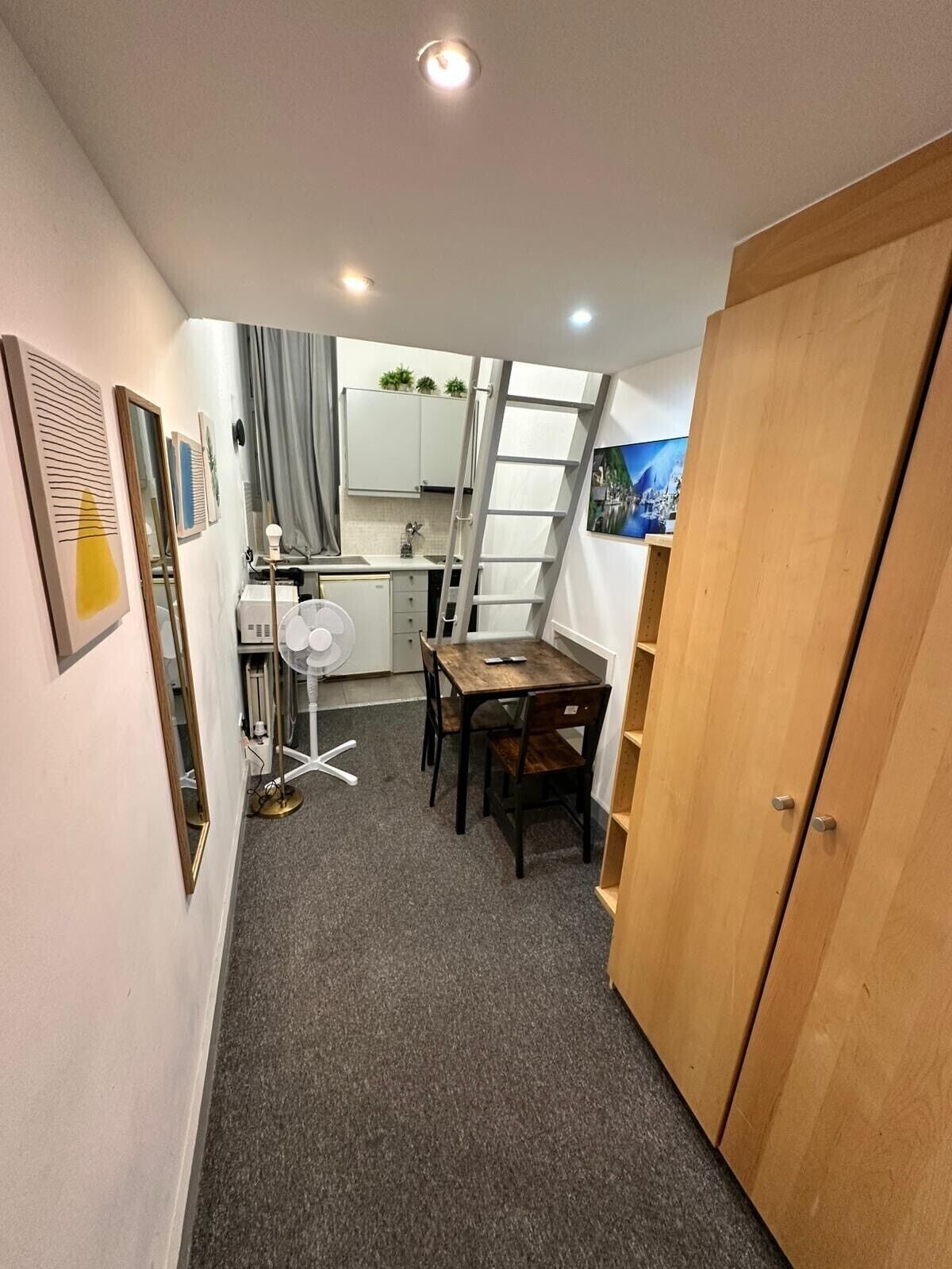 A compact room with a kitchenette, a dining area with a table and chairs, a standing fan, and a wooden wardrobe.
