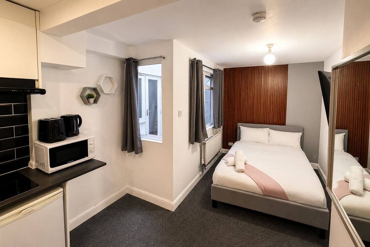 A hotel room with a bed, a microwave, a small kitchen area, a window with curtains, and a ceiling light.