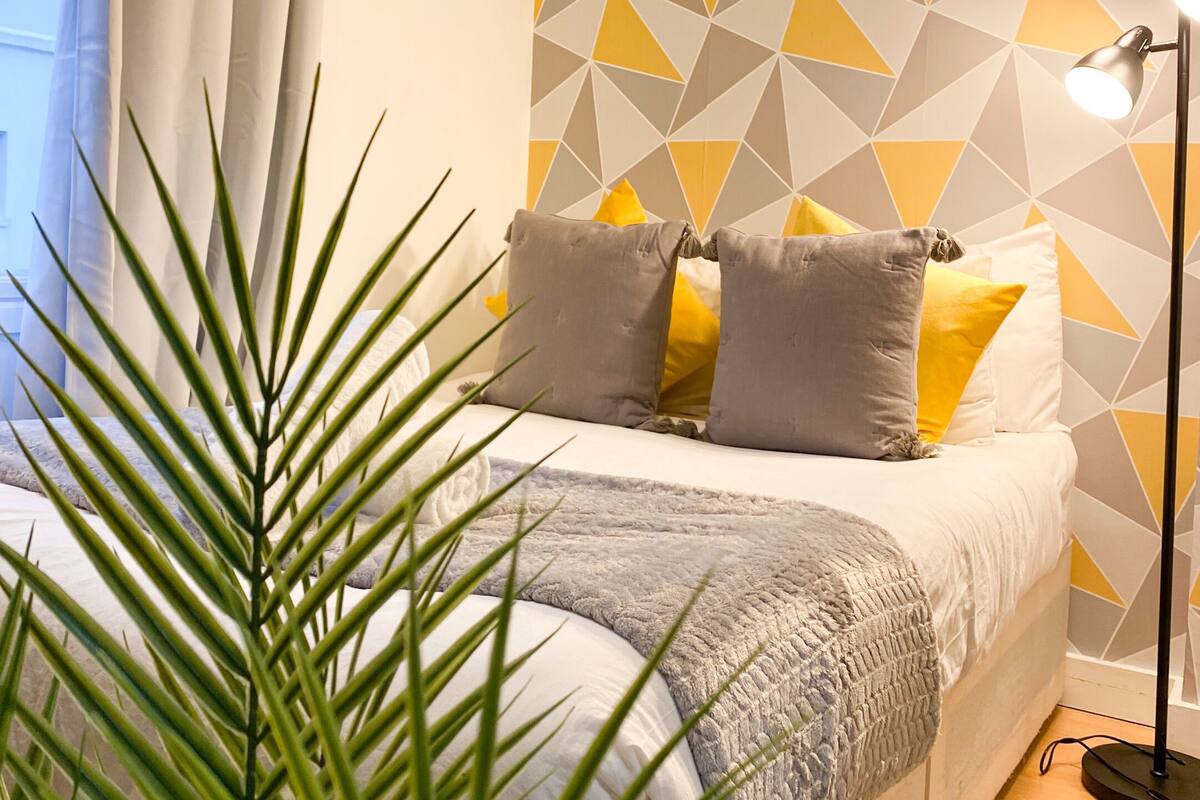 A bedroom with a bed, a geometric patterned wall, a window with curtains, a floor lamp, and a potted plant.