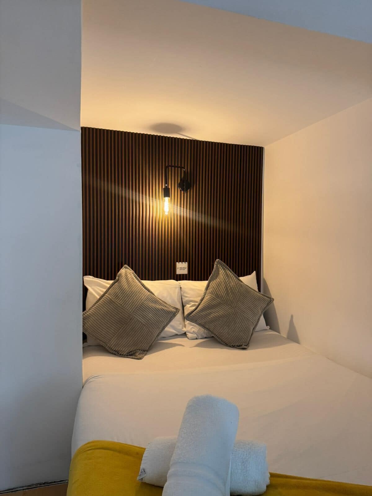 A hotel room with a bed, two pillows, a bedside lamp, and a yellow pillow on the floor.