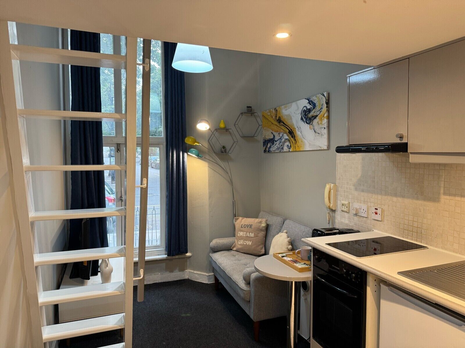 Ett kompakt kök med en spis, en diskho och ett litet matbord. En soffa och en stege syns också.