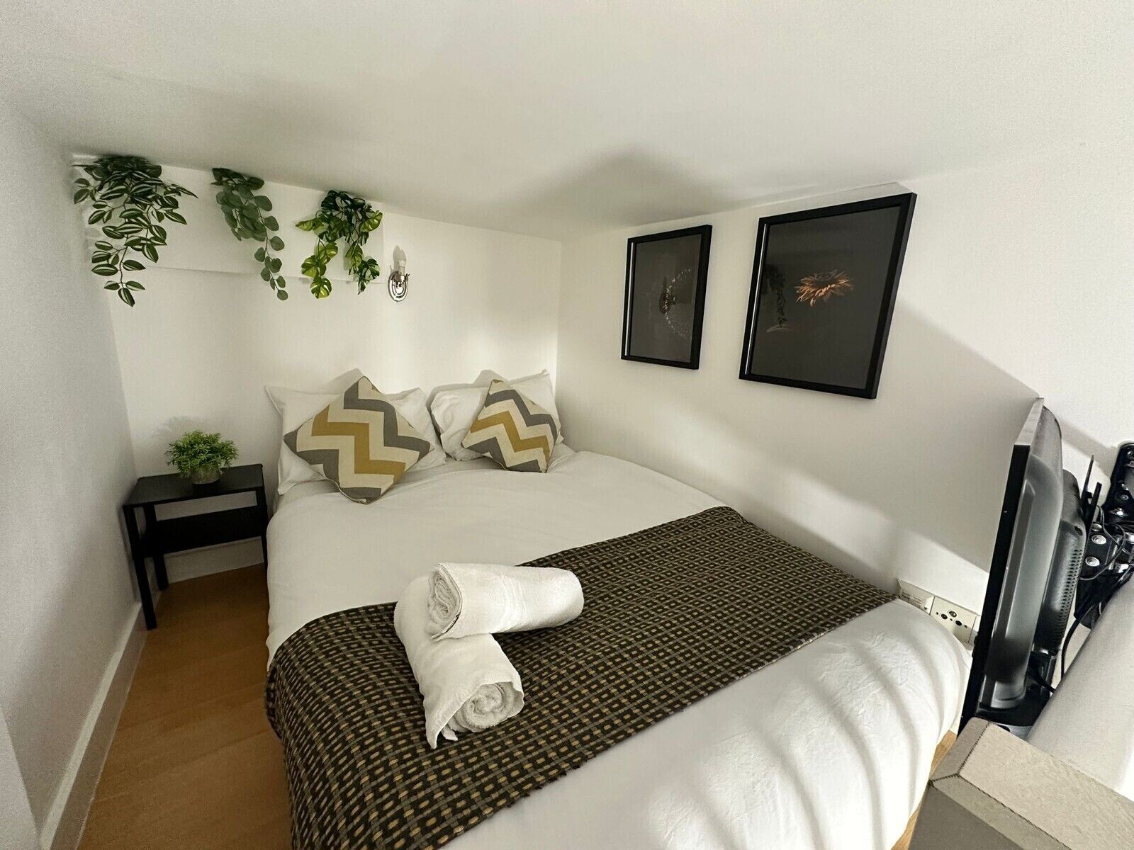 Une chambre à coucher bien rangée, avec un lit, des oreillers, une petite table de chevet agrémentée d’une plante, et deux tableaux encadrés accrochés au mur.