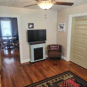 Un salon avec une cheminée, un téléviseur, un ventilateur de plafond et une porte dotée d’un porte-manteau.