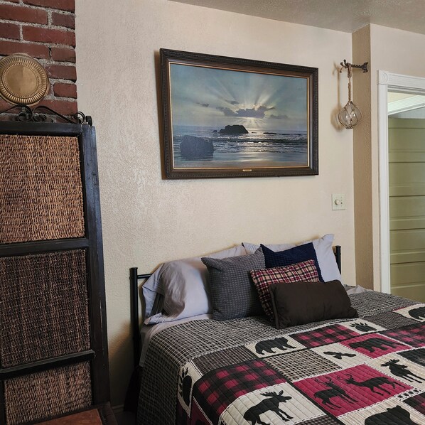 A room with a bed, a framed painting of a seascape, a wicker basket, and a door.