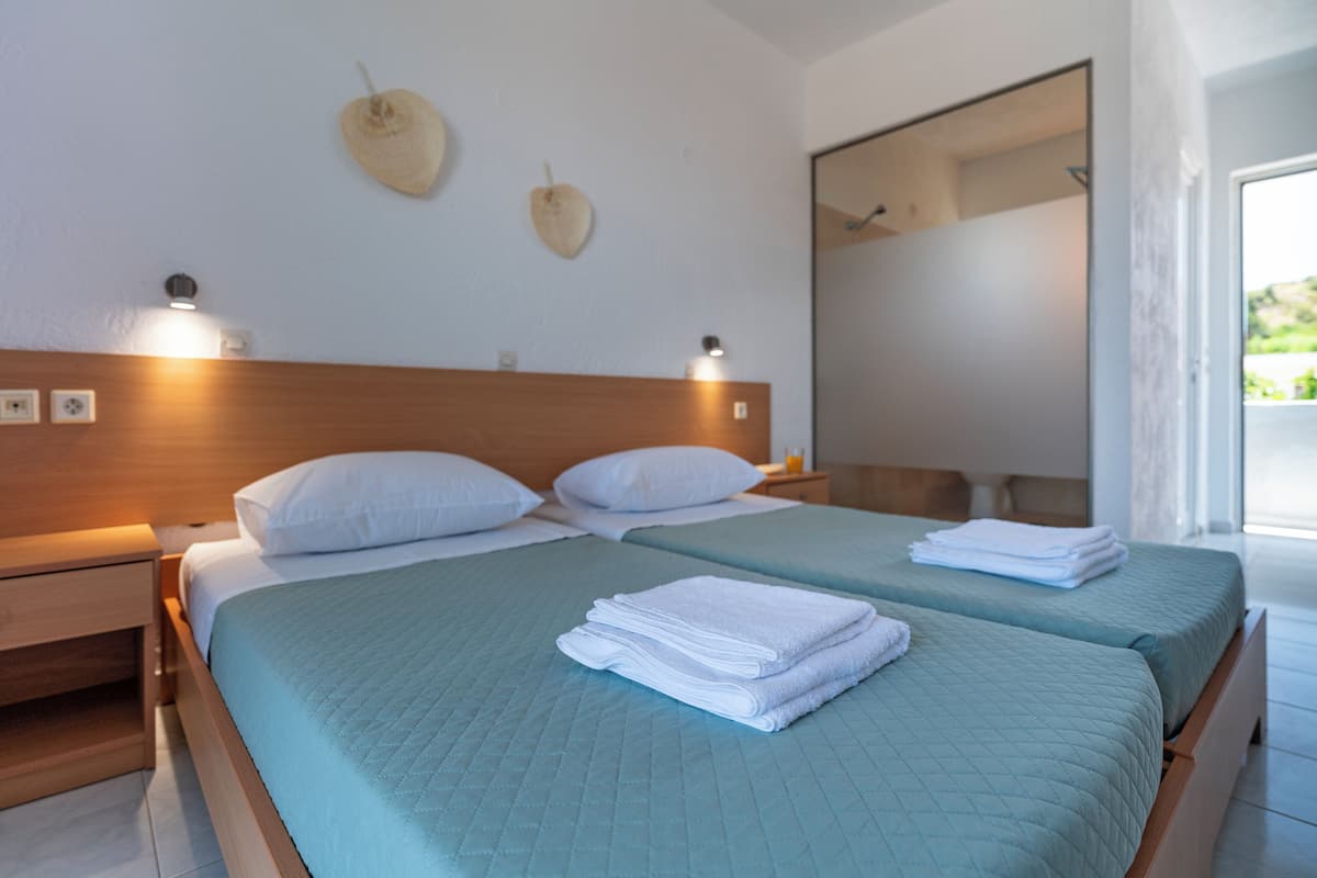 Quarto de hotel com duas camas, cada uma com colcha azul acolchoada e almofadas brancas. Há duas luminárias fixadas na parede, uma de cada lado da cabeceira.