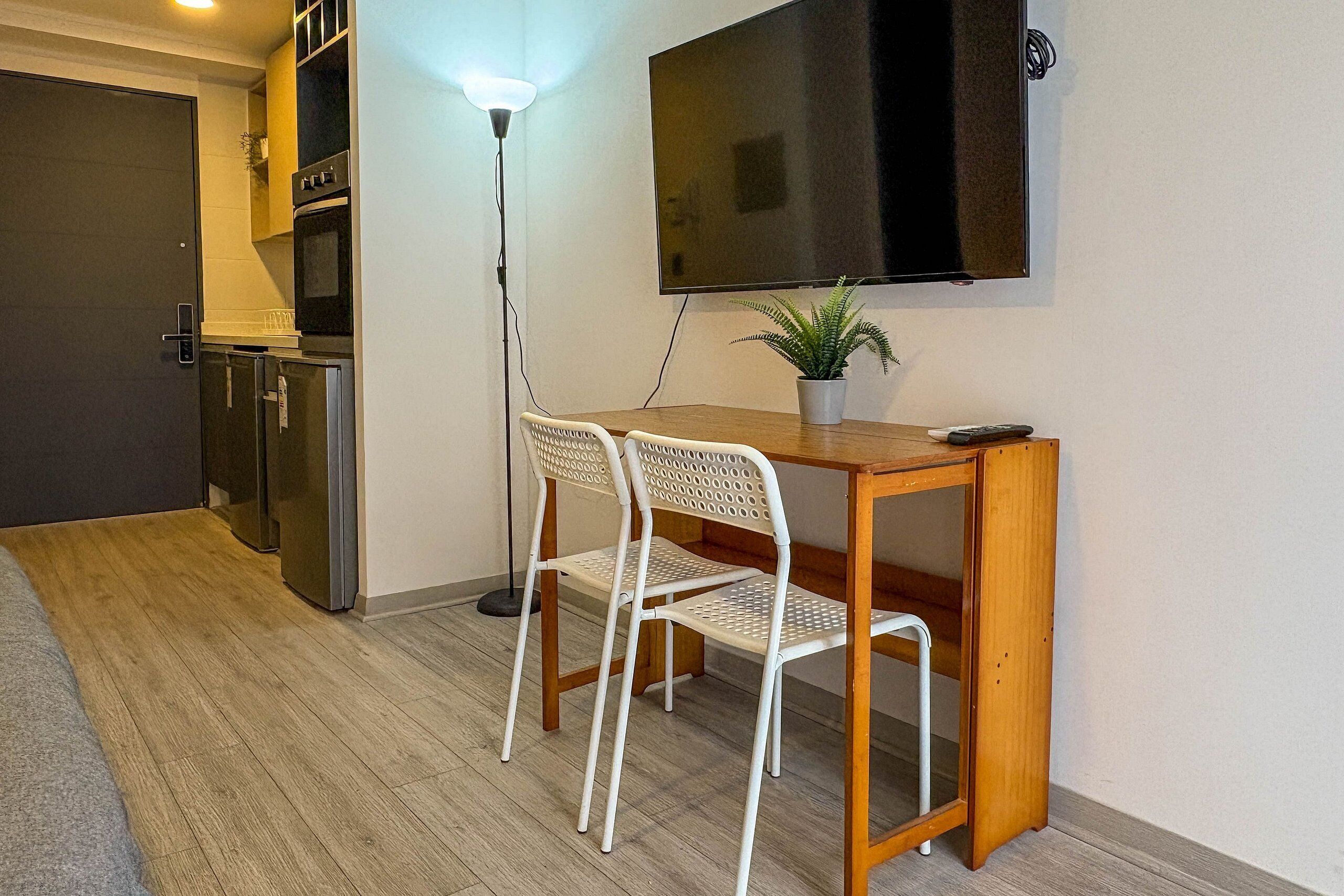 A compact kitchenette with a refrigerator, oven, and microwave. A wooden table with two white chairs and a potted plant is positioned in front of a flat-screen TV mounted on the wall.