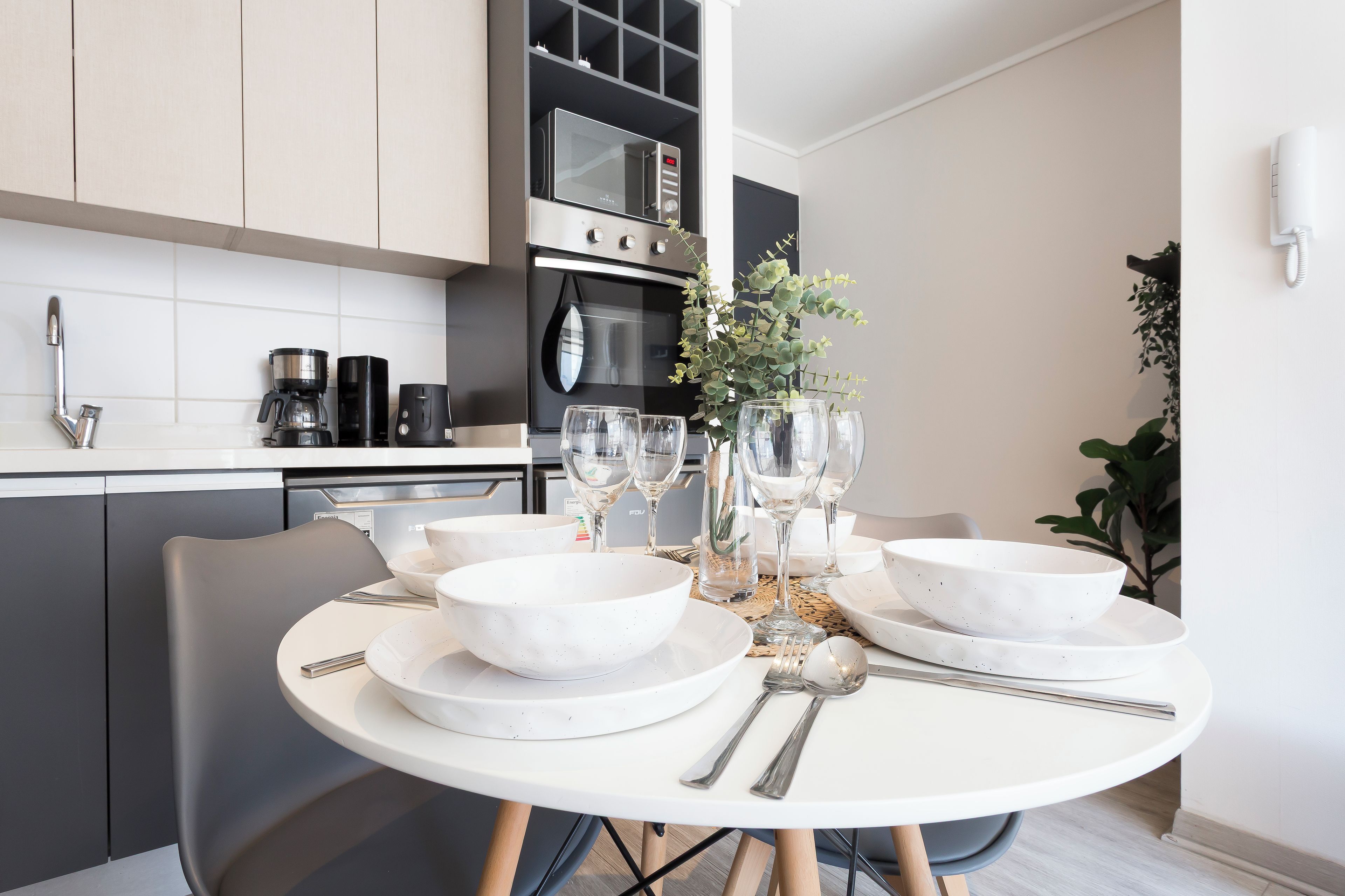 A modern kitchen with white cabinets, a built-in oven, and a dining area set with white bowls and wine glasses.
