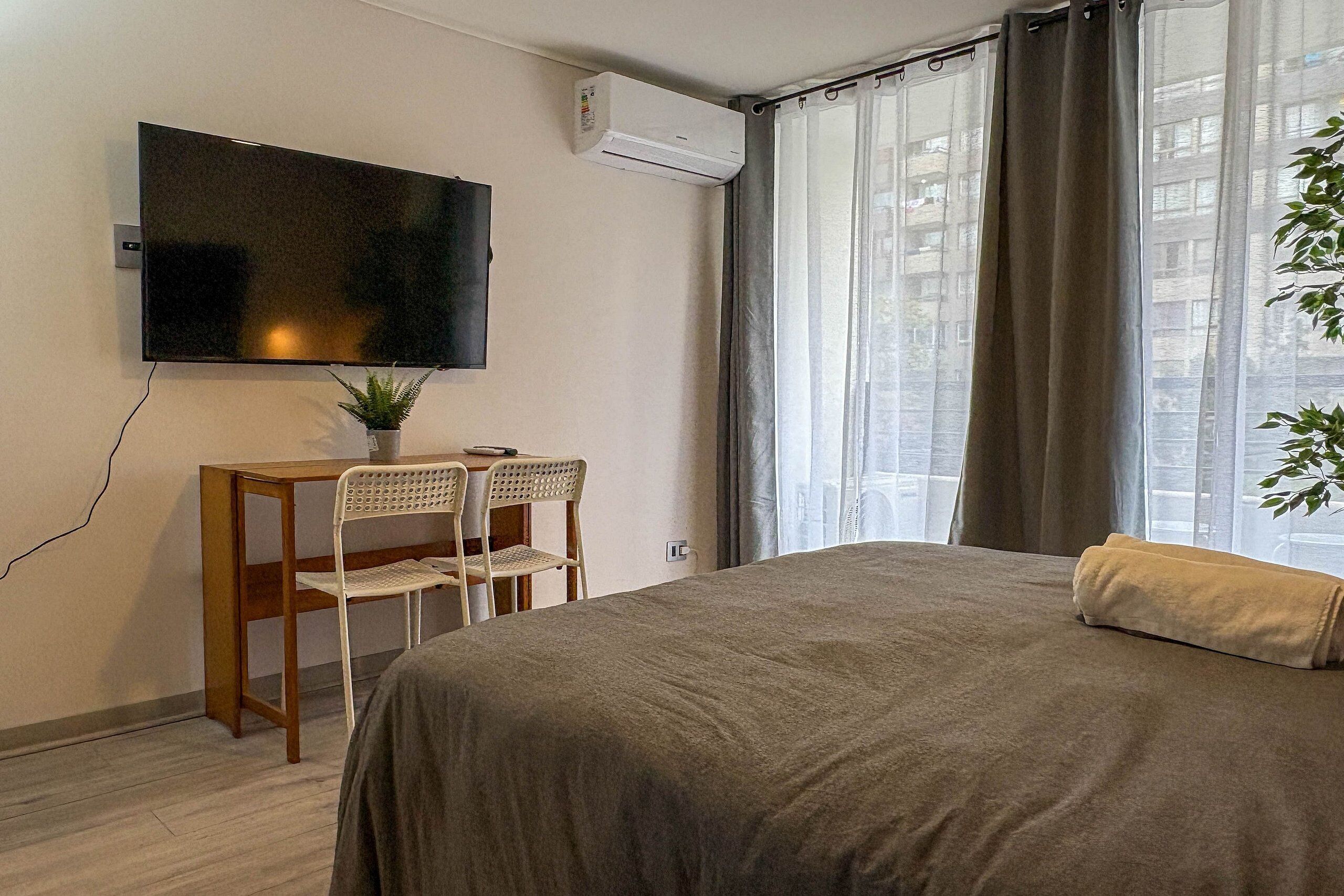 A bedroom with a bed, a TV mounted on the wall, a desk with chairs, and a window with curtains.