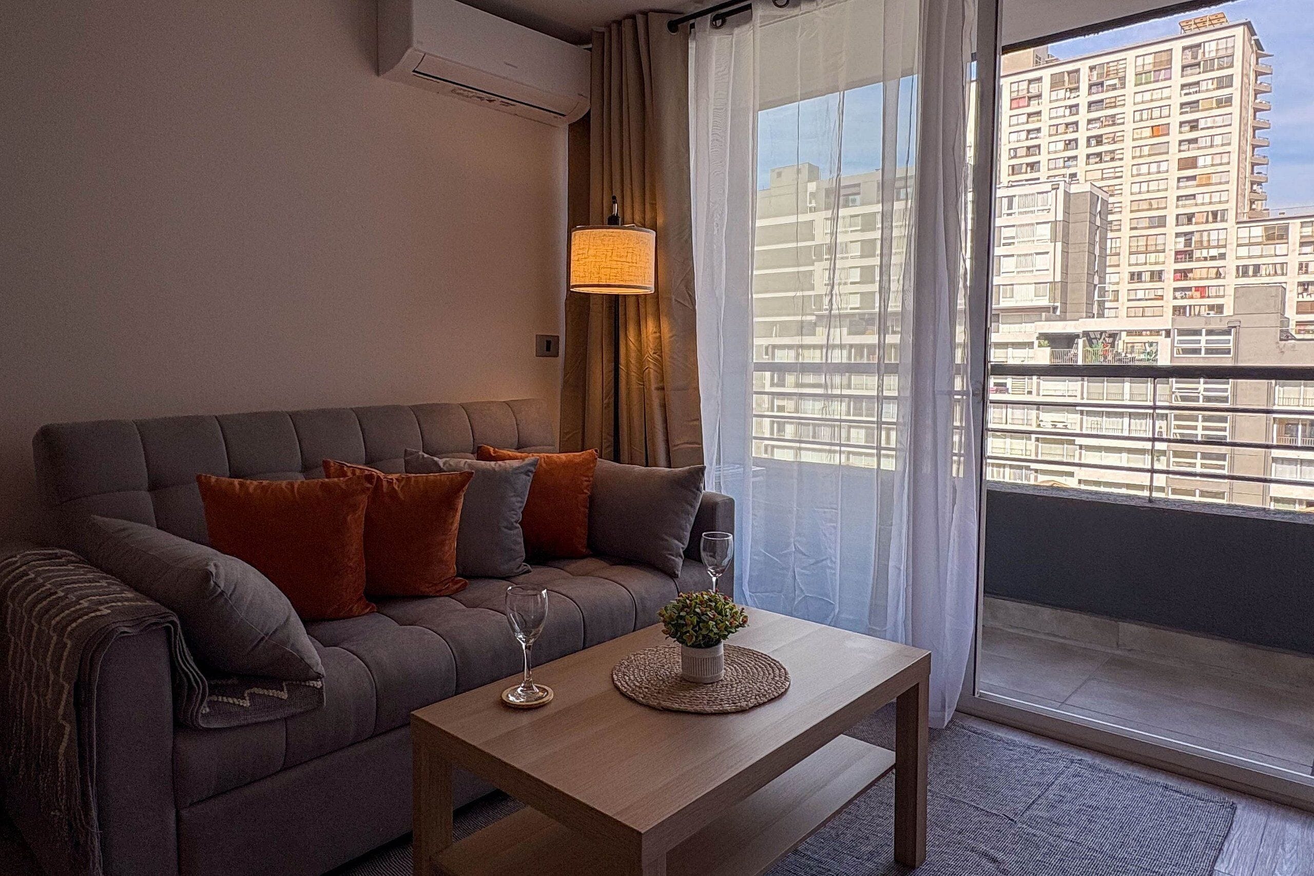 A modern living room with a grey sofa, a wooden coffee table, and a view of a high-rise building through a large window.