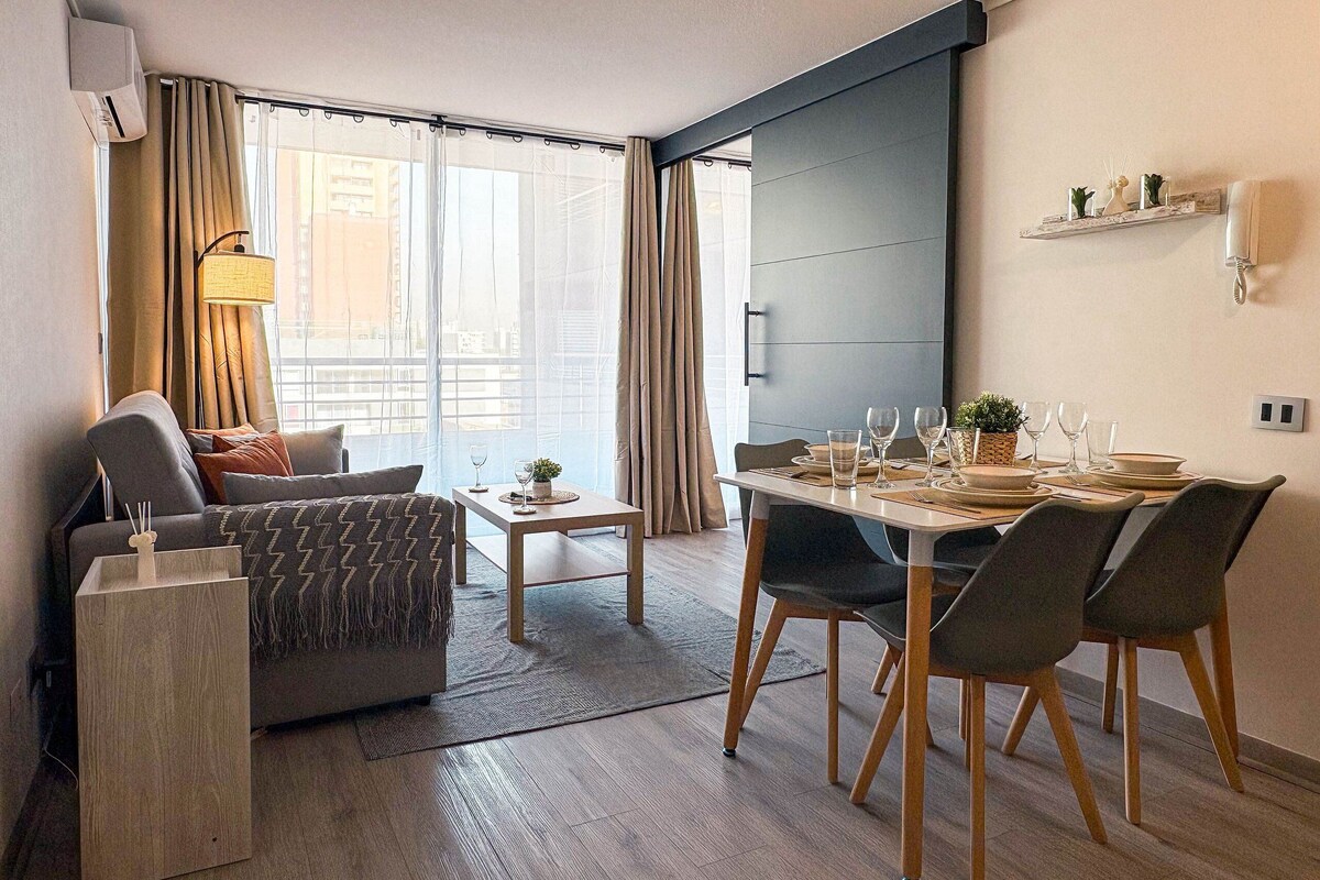 A modern living room with a sofa, dining table, and chairs. There is a small table with a lamp and a plant. The room has large windows with curtains.