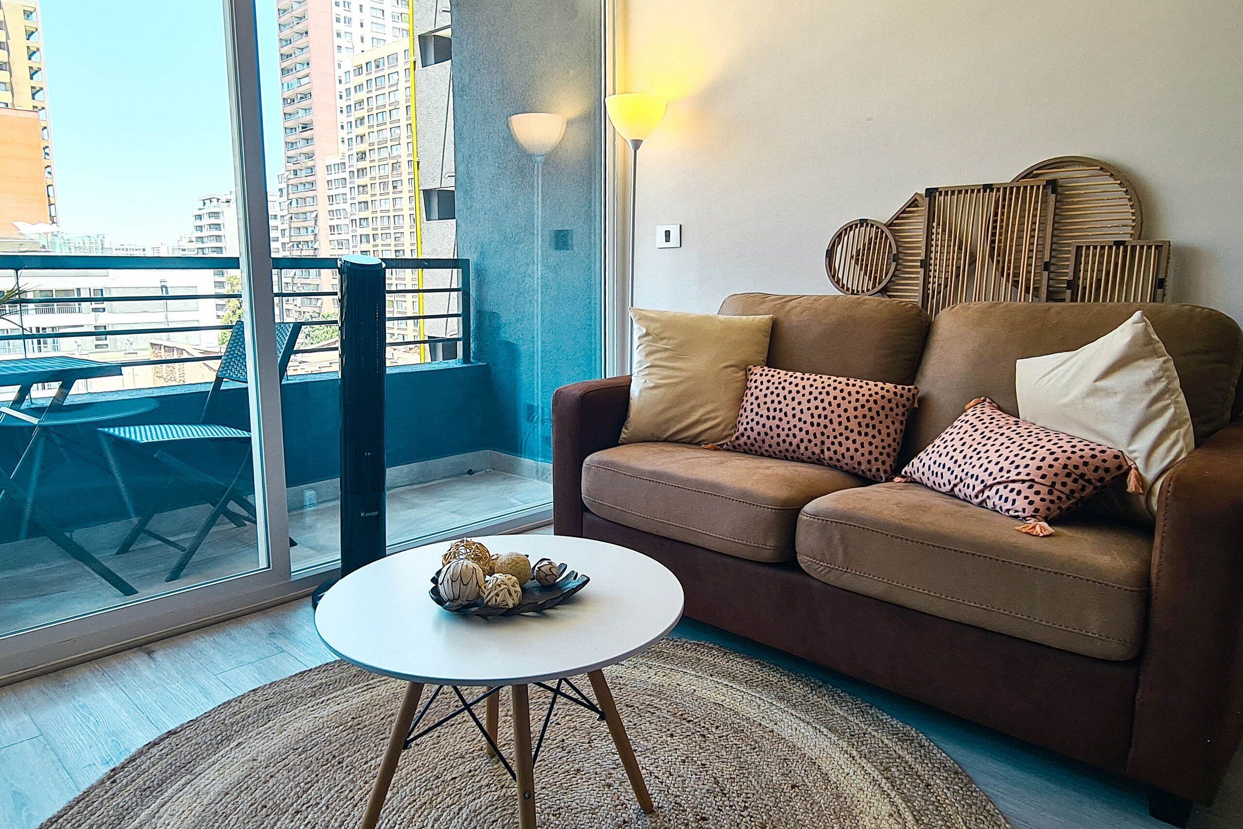 A modern living room with a brown sofa, a round white coffee table, and a view of a balcony with a cityscape.