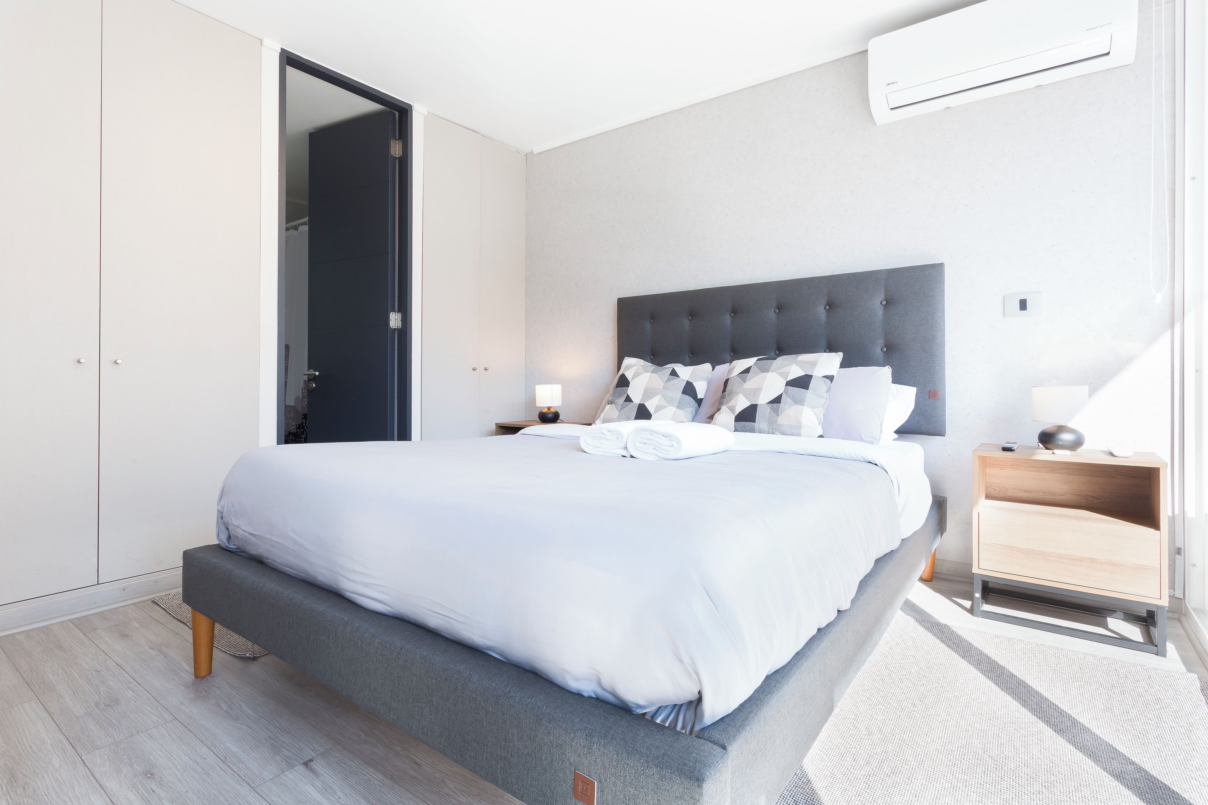 A modern bedroom with a large bed, a wardrobe, a bedside table, and an air conditioning unit.