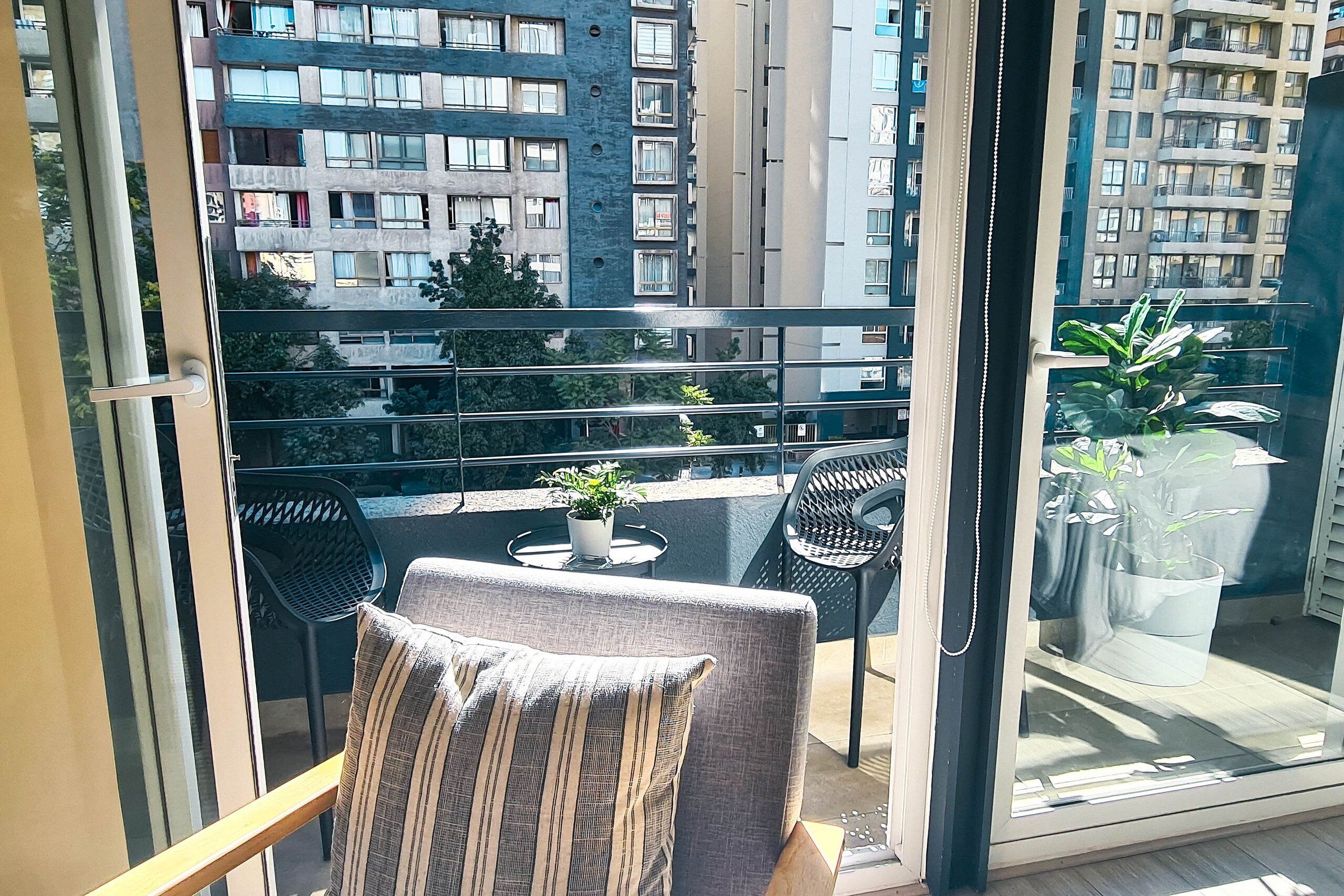 Un balcón con vistas a edificios altos, una mesa pequeña con una planta en maceta y dos sillas.