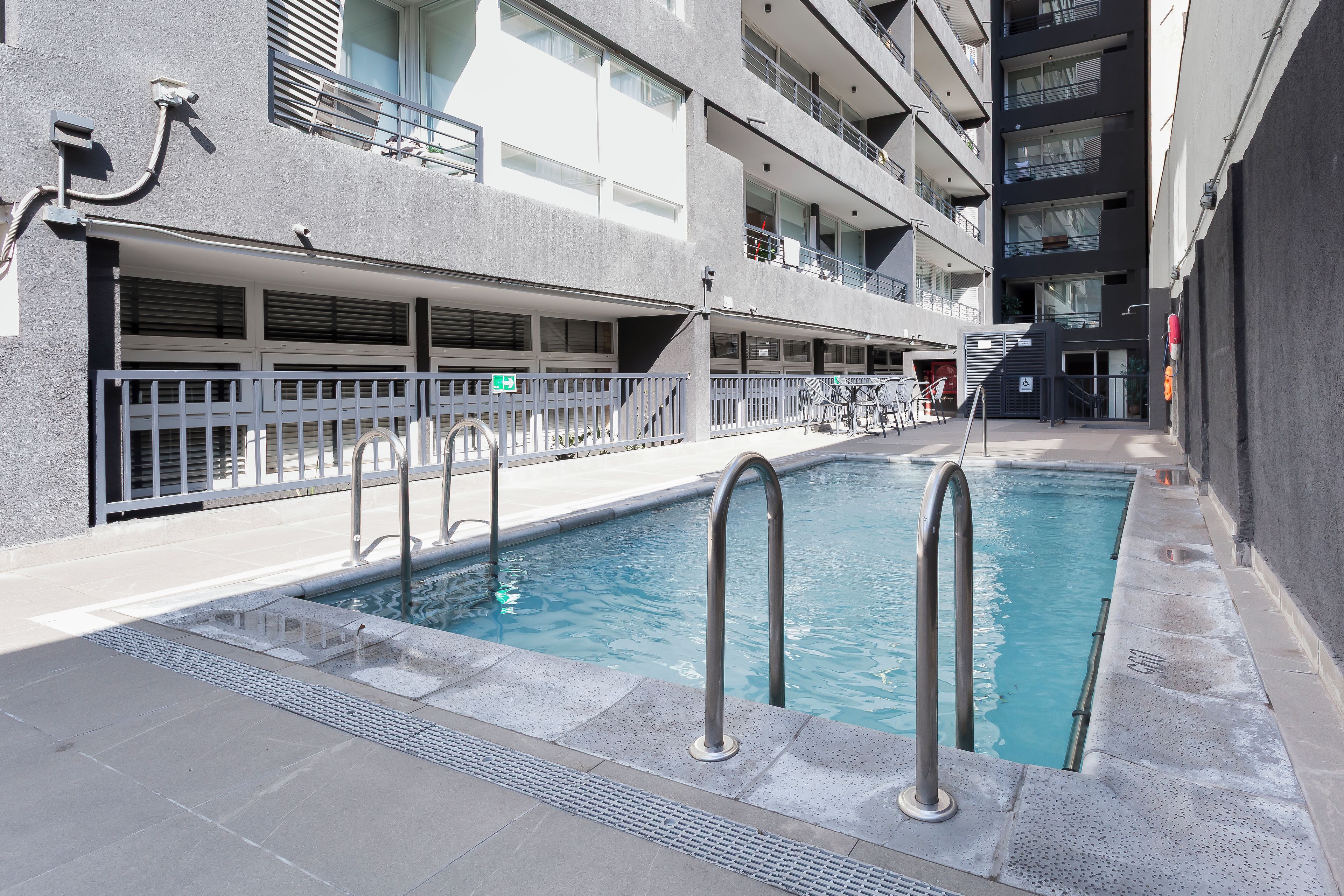 Une piscine entourée de rampes métalliques, un espace de repos couvert et un bâtiment doté de plusieurs balcons.