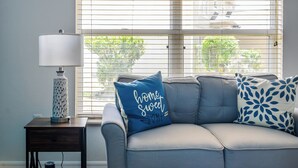 A living room with a grey sofa, a side table with a lamp, and a window with blinds. - Disney Castle Cove - Private Pool & Community Boat Ramp close to Legoland (Winter Haven)