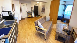 A modern hotel room with a wooden floor, a sofa, a small table, a microwave, a sink, and a door leading to another room. - 1minute walk to station, direct bus to Haneda Airport! Easy access! (大田区)