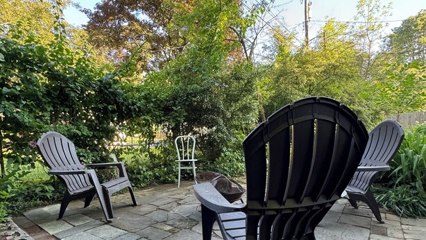 An outdoor patio with two Adirondack chairs, a small table, and a garden area with plants and a brick planter. - Walk Anywhere PSU! 3BR home in College Heights.  Summer discount now! (State College)