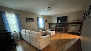 A living room with a sofa, ottoman, wooden flooring, a fireplace, and a television. - Walk Anywhere PSU! 3BR home in College Heights.  Summer discount now! (State College)