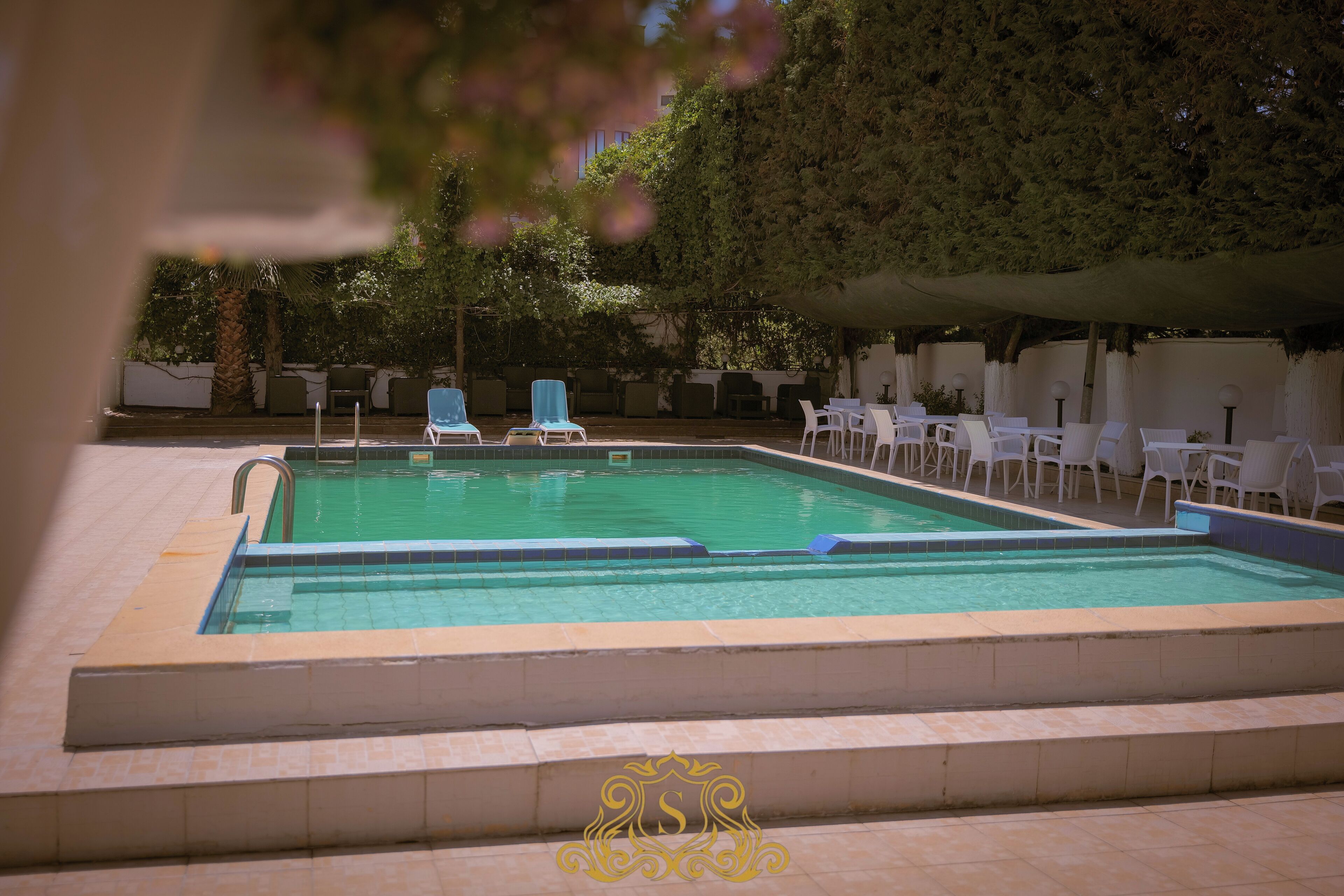 A swimming pool with lounge chairs and tables, surrounded by trees and a covered area.